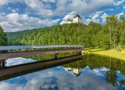 Spiegelung einer Burg auf einem Hügel im Wasser mit Holzbrücke